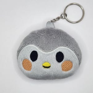 May include: A gray plush penguin keychain with black eyes, a yellow beak, and tan cheeks. The penguin has a fluffy texture and is attached to a silver metal keyring and chain. The keychain is designed to be a cute accessory.