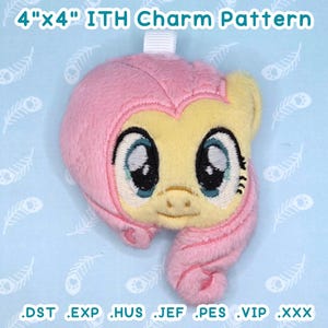 May include: A plush charm in the shape of a cartoon pony's head, primarily pink and yellow, with large blue eyes. The charm is approximately 4 inches by 4 inches and has a white loop at the top. The text "4"x4" ITH Charm Pattern" is above the charm.