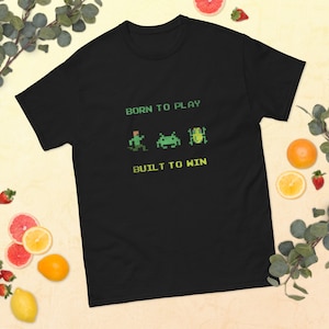 Könnte beinhalten: Schwarzes T-Shirt mit dem Text "Born to Play" und "Built to Win" in Gelb. Zwischen den Texten befinden sich drei pixelartige grüne Videospielfiguren.