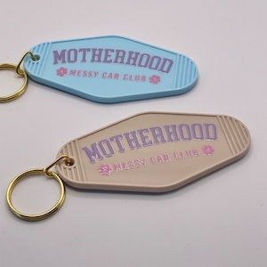 以下が含まれることがあります： 薄い青とベージュのタグが付いたキーホルダーが2つ。両方のタグには「MOTHERHOOD MESSY CAR CLUB」という文字と、文字の両側にピンクの花が描かれています。文字は紫色の色です。