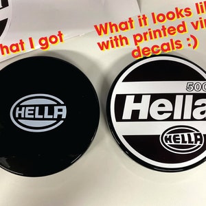 May include: Two round black car headlight covers with white Hella logos. One cover has the text "500" printed above the logo. The image shows the difference between a printed vinyl decal and a real logo.