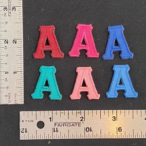 May include: Six felt letter patches in different colors. The patches are all the letter "A" in a blocky font. The colors are red, pink, blue, teal, light pink, and dark blue.