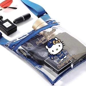 May include: Clear crossbody bag with blue trim and strap, containing cosmetics, a wallet, and a Hello Kitty graphic. Red sunglasses and a black perfume bottle are visible.