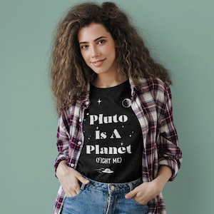 May include: A woman wearing a black t-shirt with white text that reads "Pluto is a Planet (Fight Me)". She is also wearing a plaid shirt and blue jeans.