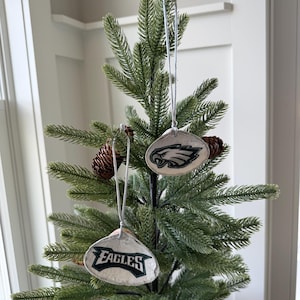 May include: Two silver-colored, oval-shaped Christmas ornaments with the Philadelphia Eagles logo in green, black, and white hang from a small artificial Christmas tree. The ornaments are suspended by silver strings. Pinecones are also visible.
