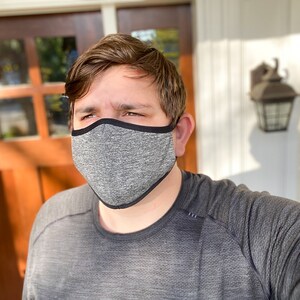 May include: A person wearing a gray face mask with a black border. The mask is made of a soft, breathable fabric. The person is standing in front of a door.
