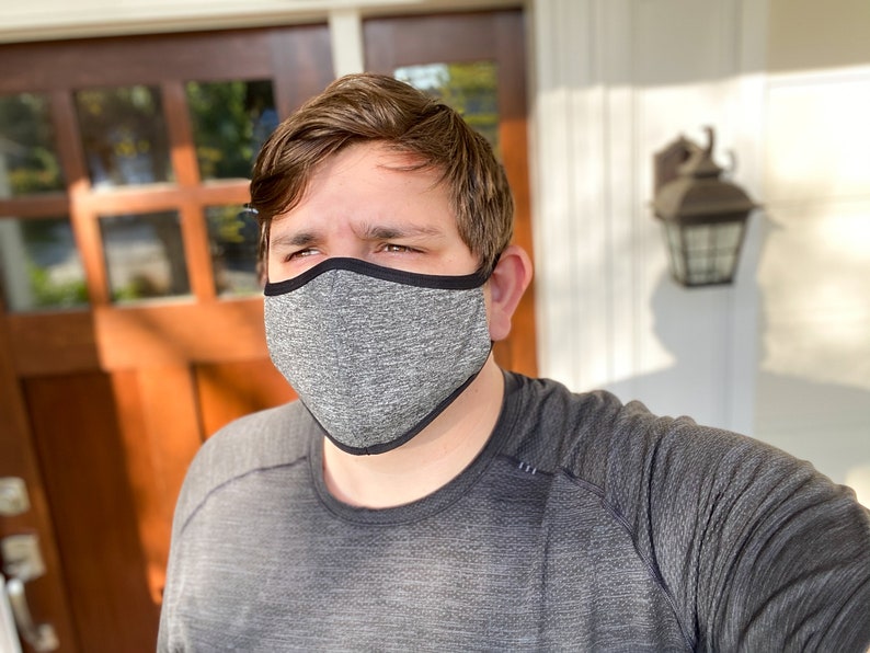 May include: A person wearing a gray face mask with a black border. The mask is made of a soft, breathable fabric. The person is standing in front of a door.