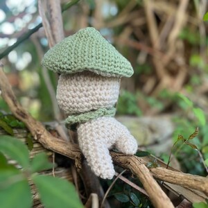 May include: A crocheted amigurumi toy shaped like a mushroom with a green hat and a beige body. The toy is sitting on a branch.