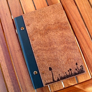 May include: A wooden journal with a black leather spine and a mushroom design burned into the front cover. The journal has brass rivets along the spine.