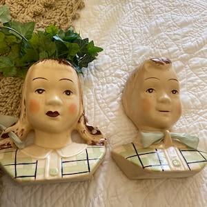 Könnte beinhalten: Zwei Keramik-Pflanztopfköpfe, einer weiblich und einer männlich, mit grün-weißem Karomuster. Beide Köpfe haben gemaltes braunes Haar und Sommersprossen.