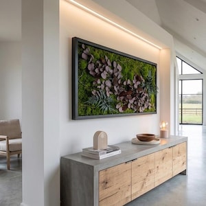 May include: A modern interior scene featuring a large framed moss art piece with burgundy and green foliage. Below, a gray and wood cabinet displays decorative items. The room has a neutral color palette with natural light.