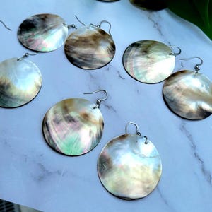 May include: A selection of round, iridescent earrings with silver hooks. The earrings have a natural, shell-like appearance with a mix of white, brown, and green hues. The earrings are presented on a white and grey surface.