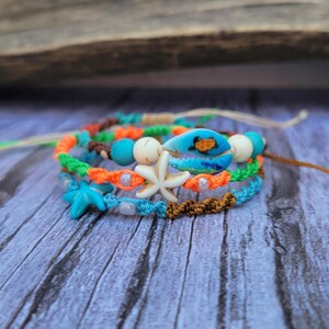 Peut inclure: Plusieurs bracelets en macramé faits à la main sur le thème de la plage. Les bracelets présentent des cordons turquoise, orange et marron, ornés d'étoiles de mer, de crabes et de perles. Les bracelets sont présentés sur une surface en bois vieilli.