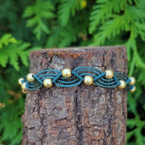 May include: A green and gold beaded bracelet with a woven design. The bracelet is displayed on a tree stump.