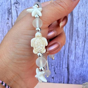 May include: A beaded bracelet featuring white starfish and turtle charms, frosted glass beads, and silver-colored accents. The bracelet is strung on a clear cord, creating a beachy, nautical theme.