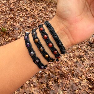 May include: A collection of black macrame bracelets adorned with colorful gemstones. The bracelets feature various shades of purple, brown, and red stones, creating a vibrant contrast against the dark cords. The bracelets are displayed on a wrist.