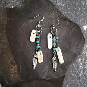 May include: A pair of dangle earrings with a bohemian style. Each earring features a silver-tone hook, a chain, turquoise beads, and a small, silver feather charm. Rectangular, off-white shell pieces are also included.