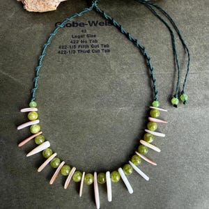 May include: A handmade necklace featuring green beads and white and pink shell accents. The necklace is strung on a dark green cord. The text "Gobe-Weis 62 Legal Size" is visible in the background.