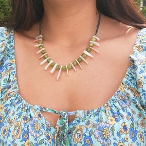 May include: A handmade necklace featuring green beads and white shell accents. The necklace is strung on a dark green cord and is worn around the neck. The necklace is a unique piece of jewelry.
