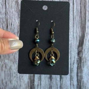 May include: A pair of dangle earrings with a celestial theme. Each earring features a brass-colored crescent moon charm, a faceted green crystal, and dark blue beads. The earrings are displayed on a black card.