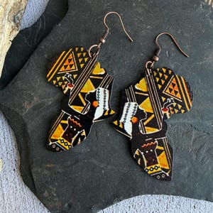 Puede incluir: Un par de pendientes de madera con forma de continente africano. Los pendientes presentan un fondo negro con patrones geométricos amarillos y naranjas y la silueta de una persona. Los pendientes tienen ganchos de color cobre.