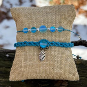 May include: Two handmade bracelets displayed on a burlap cushion. The top bracelet features blue glass beads and silver accents. The bottom bracelet is turquoise macrame with a blue bead and a silver leaf charm. The bracelets are on a natural burlap cushion.