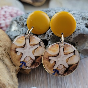 Peut inclure: Une paire de boucles d'oreilles pendantes jaune moutarde avec un charme rond en résine transparente représentant une étoile de mer et des coquillages. Les breloques sont attachées aux boucles d'oreilles avec des crochets argentés.