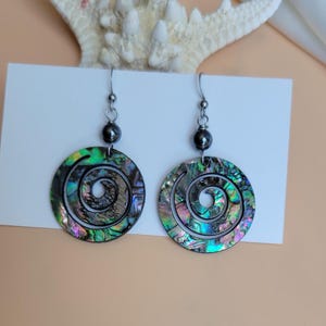 May include: A pair of iridescent abalone shell earrings with a spiral design. Each earring features a dark gray bead and a silver-tone hook. The earrings display a range of colors, including green, blue, and purple.