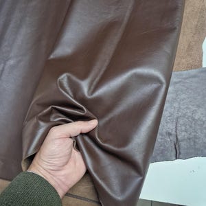 Peut inclure: Gros plan sur un tissu en cuir marron. Le tissu est lisse et a un l&eacute;ger brillant. Le tissu est tenu par une main.