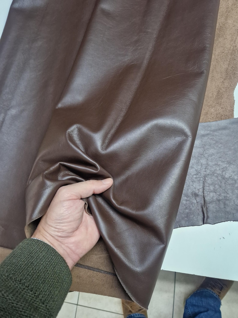 Peut inclure: Gros plan sur un tissu en cuir marron. Le tissu est lisse et a un l&eacute;ger brillant. Le tissu est tenu par une main.