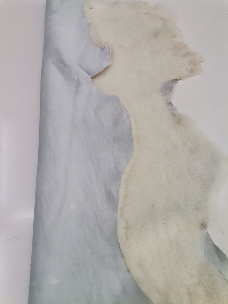 May include: A white sheepskin with a light blue leather backing. The sheepskin is folded in half, showing both the fur and the leather.