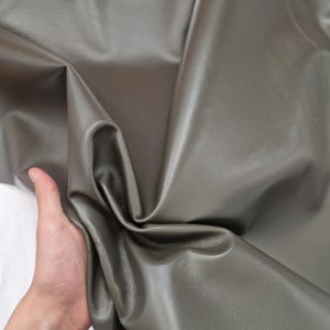 May include: A close-up of a sheet of olive-green leather. The leather is folded and draped, revealing its smooth texture and subtle sheen. The material is held by a hand, showing the flexibility of the leather. The background is white.