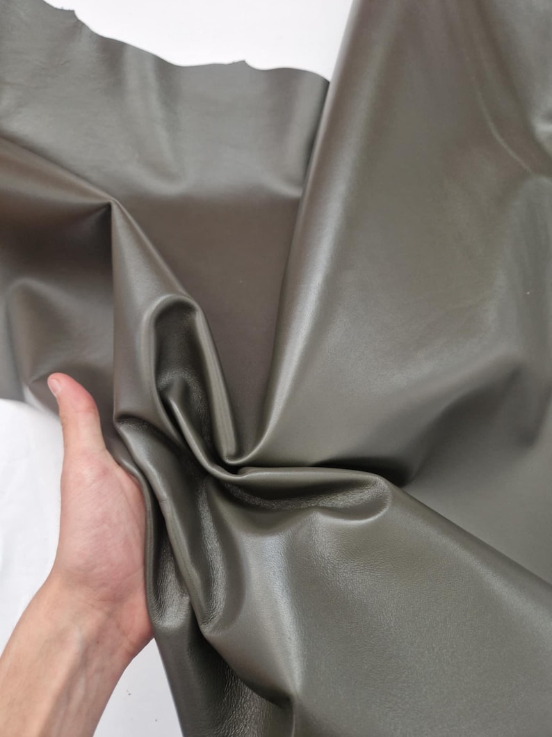May include: A close-up of a sheet of olive-green leather. The leather is folded and draped, revealing its smooth texture and subtle sheen. The material is held by a hand, showing the flexibility of the leather. The background is white.