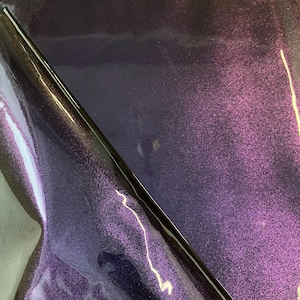 May include: A sheet of iridescent purple vinyl with a glossy finish. The material has a speckled texture, with hints of pink and blue. The vinyl is partially rolled, revealing its flexible nature.