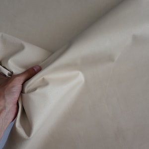 K&ouml;nnte beinhalten: Nahaufnahme eines beigefarbenen Stoffes, der von einer Hand gehalten wird. Der Stoff ist glatt und hat einen leichten Glanz.