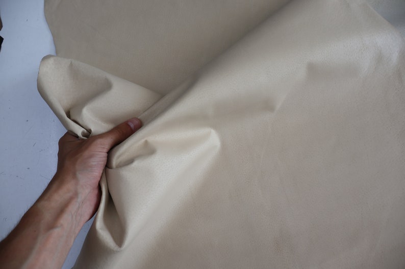 K&ouml;nnte beinhalten: Nahaufnahme eines beigefarbenen Stoffes, der von einer Hand gehalten wird. Der Stoff ist glatt und hat einen leichten Glanz.