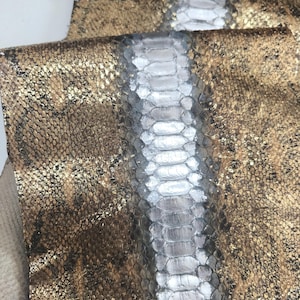 May include: Close-up of two pieces of snakeskin leather. One piece is a metallic gold color with dark brown spots. The other piece is a light gray color with a scale pattern. The leather is likely used for fashion accessories.