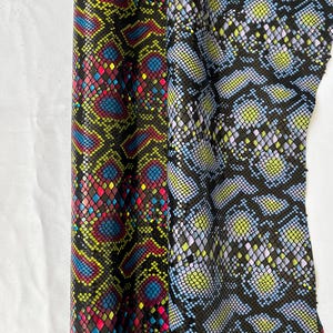 May include: Two pieces of fabric with a snakeskin pattern. One fabric is black with a bright yellow, pink, and blue pattern. The other fabric is black with a light blue and green pattern.