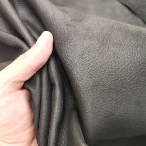 May include: Close-up of a piece of black leather with a textured surface. The leather is folded and held by a hand.