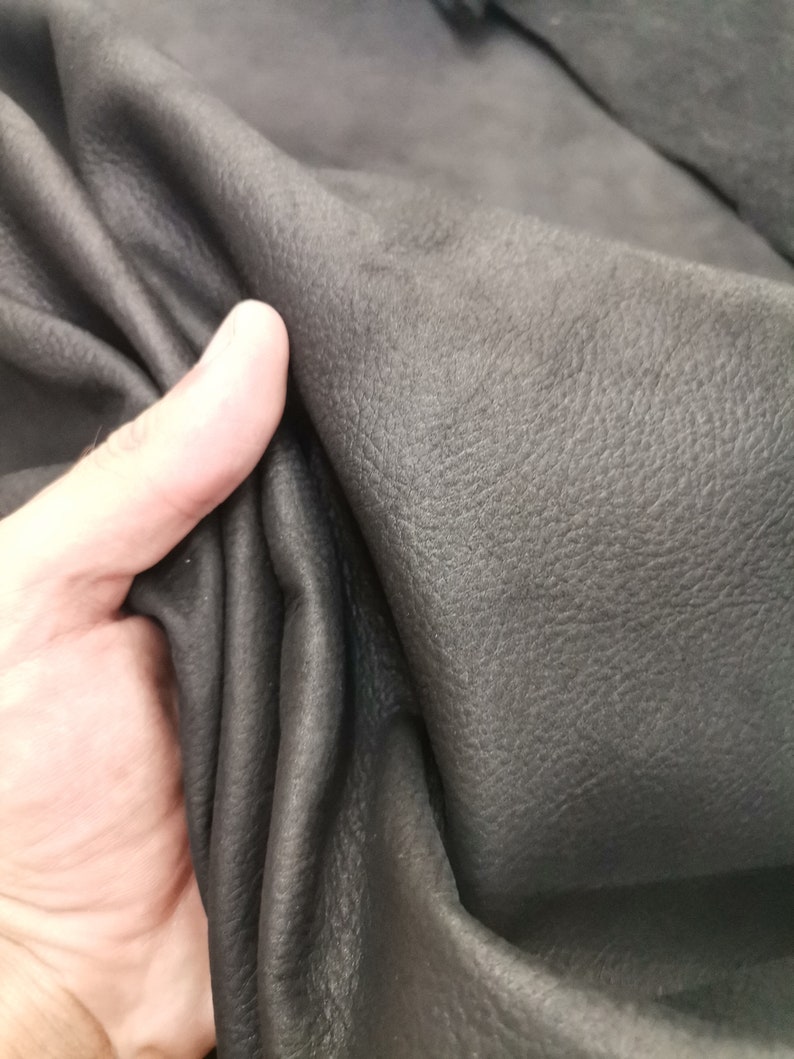 May include: Close-up of a piece of black leather with a textured surface. The leather is folded and held by a hand.
