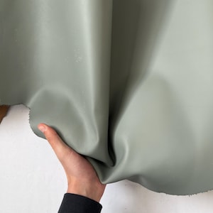 Puede incluir: Un trozo de cuero suave, flexible y de color verde. El cuero est&aacute; doblado por la mitad y est&aacute; siendo sostenido por una mano.