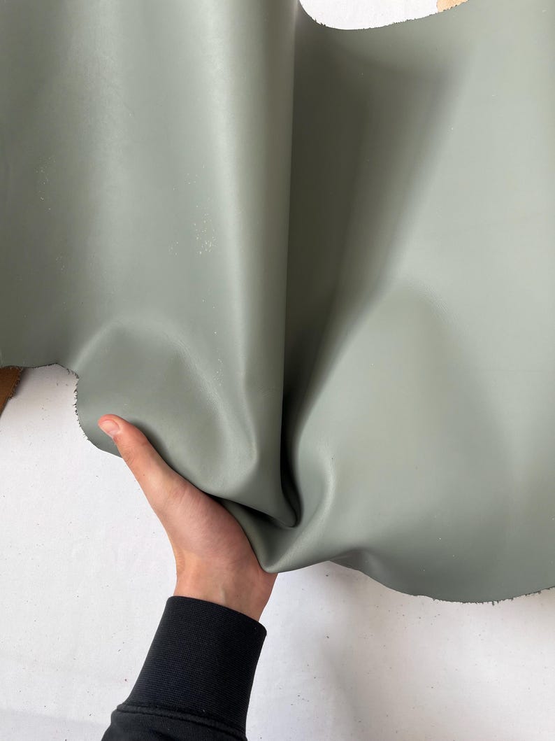 Puede incluir: Un trozo de cuero suave, flexible y de color verde. El cuero est&aacute; doblado por la mitad y est&aacute; siendo sostenido por una mano.