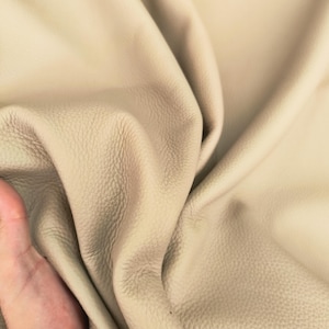 May include: Close-up of a beige leather material with a textured surface. The leather is draped in soft folds, showcasing its flexibility and natural grain. The color is a warm, neutral tone.