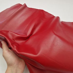 May include: A piece of red leather with a glossy finish. The leather is folded and held by a hand, showing its flexibility and texture. The material appears smooth and vibrant, suitable for various crafting projects.