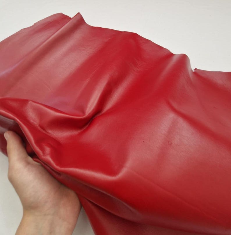 May include: A piece of red leather with a glossy finish. The leather is folded and held by a hand, showing its flexibility and texture. The material appears smooth and vibrant, suitable for various crafting projects.