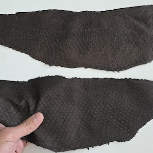 Könnte beinhalten: Zwei Stücke dunkelbraunes Leder mit einem strukturierten, schuppenartigen Muster. Ein Stück wird von einer Hand gehalten. Das Leder hat eine leicht unregelmäßige Form und scheint aus einer größeren Haut geschnitten zu sein. Der Hintergrund ist weiß.