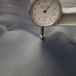 May include: A dial gauge measuring the thickness of a piece of navy blue leather. The gauge is silver with a white face and black markings, indicating a measurement of approximately 0.1 mm. The leather has a textured surface.