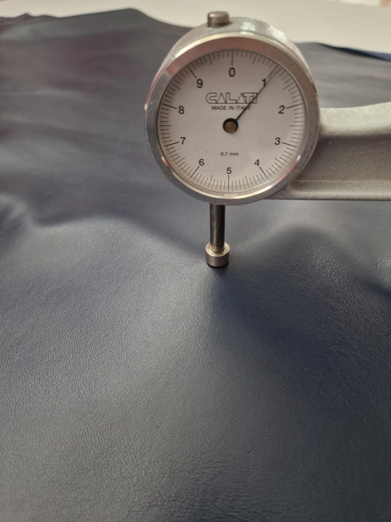 May include: A dial gauge measuring the thickness of a piece of navy blue leather. The gauge is silver with a white face and black markings, indicating a measurement of approximately 0.1 mm. The leather has a textured surface.