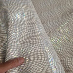 May include: Close-up of a sheet of iridescent, textured fabric. The material has a grid-like pattern with a shimmering, pearlescent finish. The fabric reflects light, creating a rainbow effect. The material is folded, showing its flexibility.
