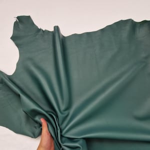 May include: A large piece of emerald green leather, with a smooth, matte finish. The leather is irregularly shaped, with natural edges. The material is held by a hand, showing the supple texture and potential for crafting. The color is consistent throughout the hide.